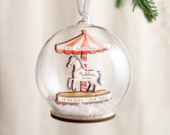 May include: A clear glass Christmas ornament with a wooden carousel inside. The carousel has a red and white striped canopy and a white horse with a blue mane and tail. The ornament also includes the text "First Christmas 2023", "Madeleine Mildred", "14.08.2023", and "6lb 10oz".