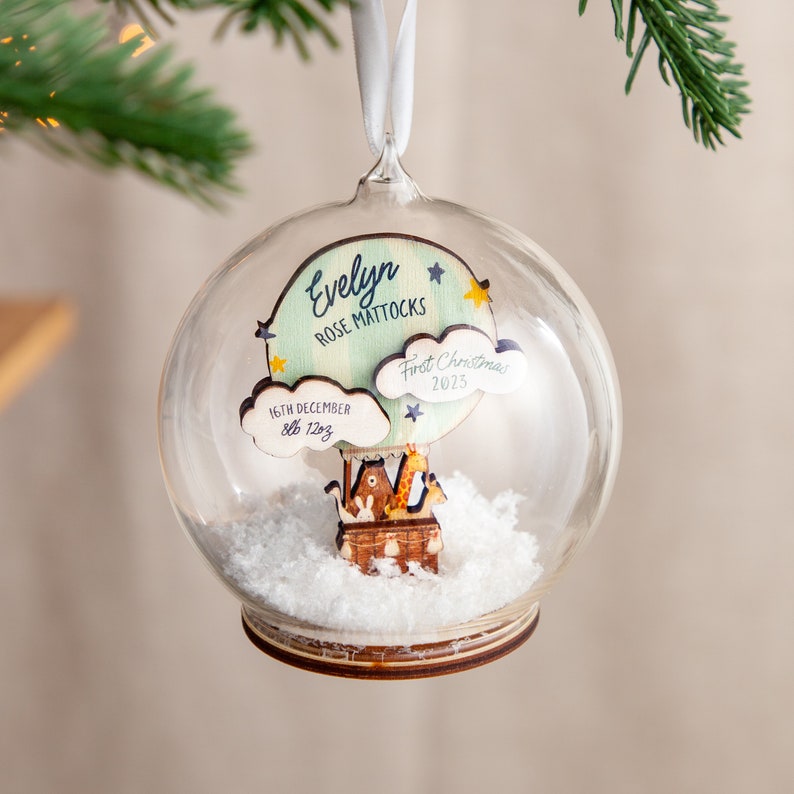 May include: A glass snow globe Christmas ornament with a wooden hot air balloon inside. The balloon has a blue and green cloud with the text "First Christmas 2023" and "16th December 8lb 12oz". The balloon is carrying a giraffe, a bunny, and a bear. The globe has a wooden base and is filled with fake snow.