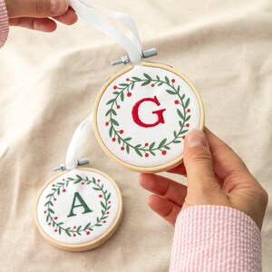 May include: Two circular wooden embroidery hoops containing white fabric with embroidered holiday wreaths. One hoop features a red letter G, and the other features a green letter A. Each hoop has a white ribbon attached for hanging purposes.
