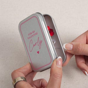 May include: A silver metal gift box with a pink border and the text "FOR MY VALENTINE Carly" in pink script. Inside the box is a small red rose. The box is being opened by a person.