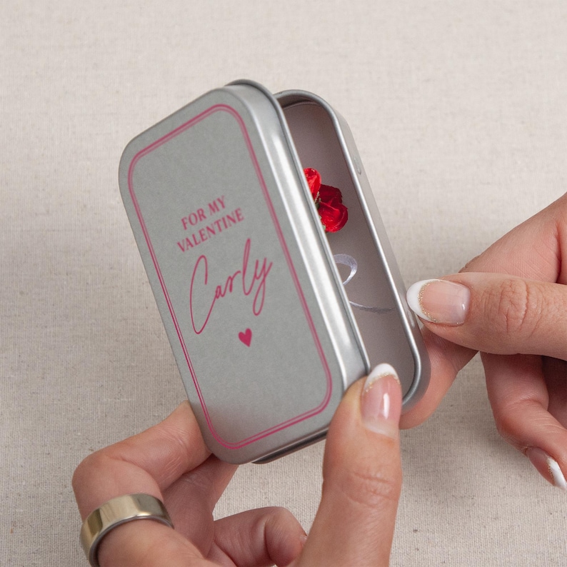 May include: A silver metal gift box with a pink border and the text "FOR MY VALENTINE Carly" in pink script. Inside the box is a small red rose. The box is being opened by a person.