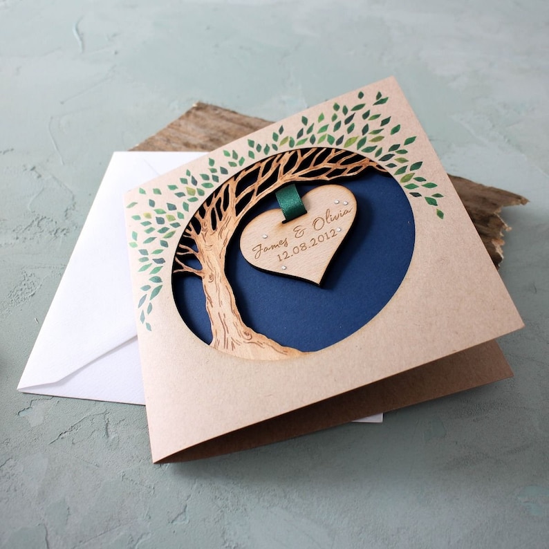 May include: A brown card with a cutout of a tree with green leaves. A wooden heart with the text "James & Olivia 12.08.2012" is hanging from the tree.