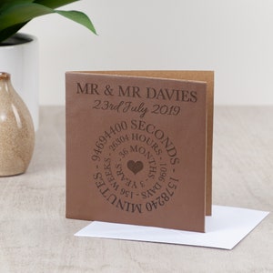 May include: A brown leather card with the text "MR & MR DAVIES 23rd July 2019" and a circle of text that reads "94694400 SECONDS - 157680 MINUTES - 26304 HOURS - 1096 DAYS - 36 MONTHS - 3 YEARS - 156 WEEKS" with a heart symbol in the center.