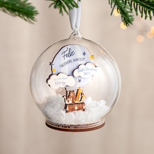 May include: A clear glass ball ornament with a wooden hot air balloon inside. The balloon has a giraffe, a bear, and a bunny on it. The ornament also has a blue cloud with the text "Felix Theodore Huntley First Christmas 2023" and another cloud with the text "21st October 3.570 kg".