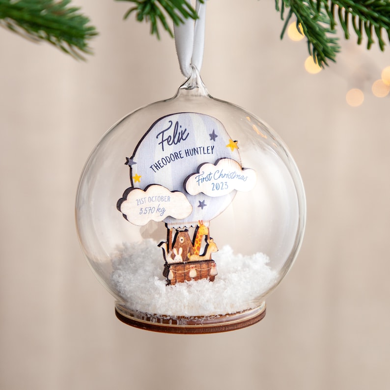 May include: A clear glass ball ornament with a wooden hot air balloon inside. The balloon has a giraffe, a bear, and a bunny on it. The ornament also has a blue cloud with the text "Felix Theodore Huntley First Christmas 2023" and another cloud with the text "21st October 3.570 kg".