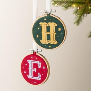 May include: Two circular embroidery hoop ornaments hanging from white ribbons. The top hoop features a dark green fabric with a gold letter H and small gold stars. The bottom hoop features a red fabric with a white letter E and small white stars.