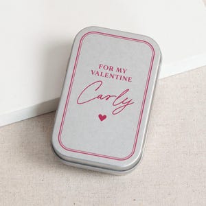 May include: A rectangular silver tin with a pink border and the words "FOR MY VALENTINE" and "Carly" in pink script. A small pink heart is below the name. The tin is on a white surface.