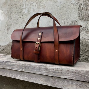 Könnte beinhalten: Eine braune Ledertasche mit einer Schließe und Messing-Akzenten. Die Tasche hat zwei obere Griffe und einen Klappverschluss.