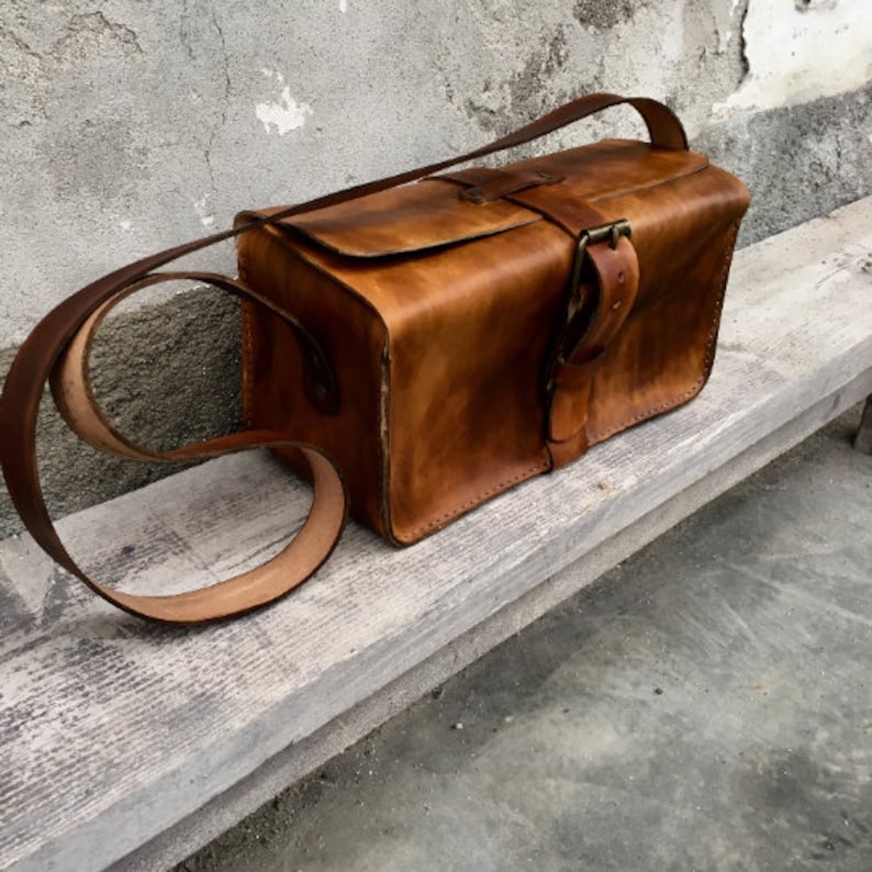 Pu&ograve; includere: Una borsa a tracolla in pelle marrone con chiusura a fibbia e una lunga tracolla. La borsa &egrave; appoggiata su una superficie di legno.