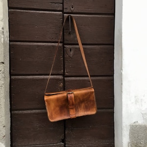 Pu&ograve; includere: Una borsa a tracolla in pelle marrone con un cinturino lungo appeso a un gancio su una porta di legno. La borsa ha una chiusura a fibbia e un aspetto usurato e invecchiato.