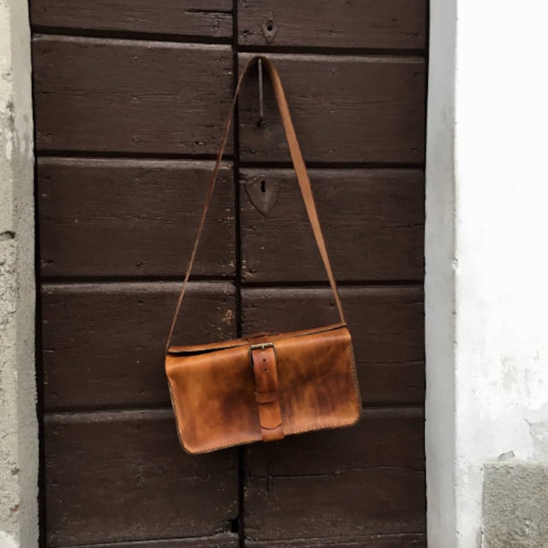 Pu&ograve; includere: Una borsa a tracolla in pelle marrone con un cinturino lungo appeso a un gancio su una porta di legno. La borsa ha una chiusura a fibbia e un aspetto usurato e invecchiato.