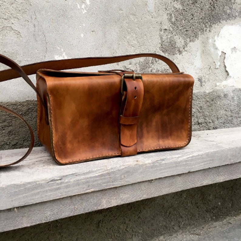Pu&ograve; includere: Una borsa a tracolla in pelle marrone con chiusura a fibbia e una lunga tracolla. La borsa &egrave; appoggiata su una superficie di legno.