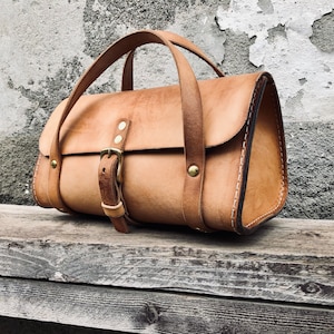 May include: A brown leather satchel bag with a brass buckle and a top handle. The bag has a flap closure and a stitched design.
