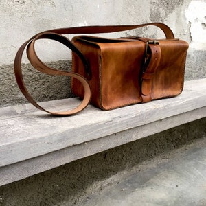 Pu&ograve; includere: Una borsa a tracolla in pelle marrone con una lunga tracolla e una chiusura a fibbia. La borsa &egrave; appoggiata su una superficie in legno grigio chiaro.