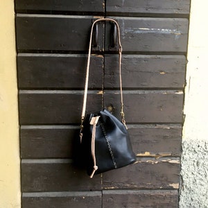 May include: A black leather bucket bag with a drawstring closure and a light pink strap. The bag has a studded design and is hanging on a wooden door.
