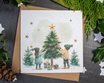 May include: A Christmas card featuring a watercolor illustration of two children decorating a Christmas tree with ornaments and presents. The card has a white background with gold glitter and stars.