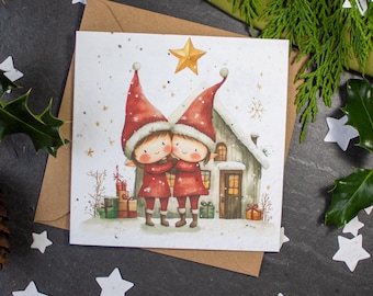 May include: A Christmas card featuring two elves in red hats and coats standing in front of a small house with a star above them. The card has a white background with a snowy scene.