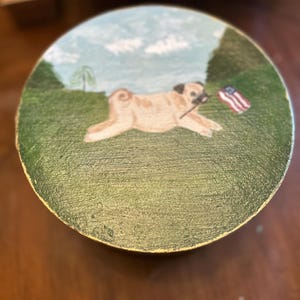 May include: A round wooden box lid featuring a hand-painted scene of a tan pug dog running across a green grassy field. The dog holds a small American flag in its mouth. The background shows a light blue sky with white clouds and green trees.