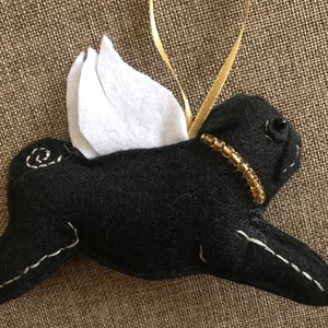May include: A black felt ornament shaped like a dog with white felt wings and gold bead collar. The dog has white stitched details and a gold ribbon for hanging. The ornament is on a textured brown surface.