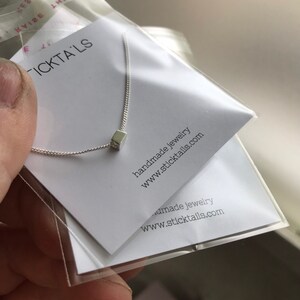 Può includere: Una collana d'argento con un piccolo ciondolo a forma di cubo. La collana è confezionata in una scatola bianca con il testo "STICKTAILS" e "gioielli fatti a mano www.sticktails.com" stampato su di essa.