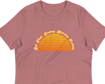May include: A mauve women's t-shirt featuring a retro-style graphic of a sunset with the text "Got That Sunday Morning Feeling" around it.