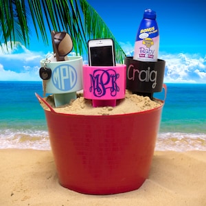 May include: Three personalized sand cup holders in a red bucket. The cup holders are blue, pink, and black. The blue cup holder has the initials "MPD" on it. The pink cup holder has the initials "MPK" on it. The black cup holder has the name "Craig" on it. The cup holders are sitting in a bucket of sand.