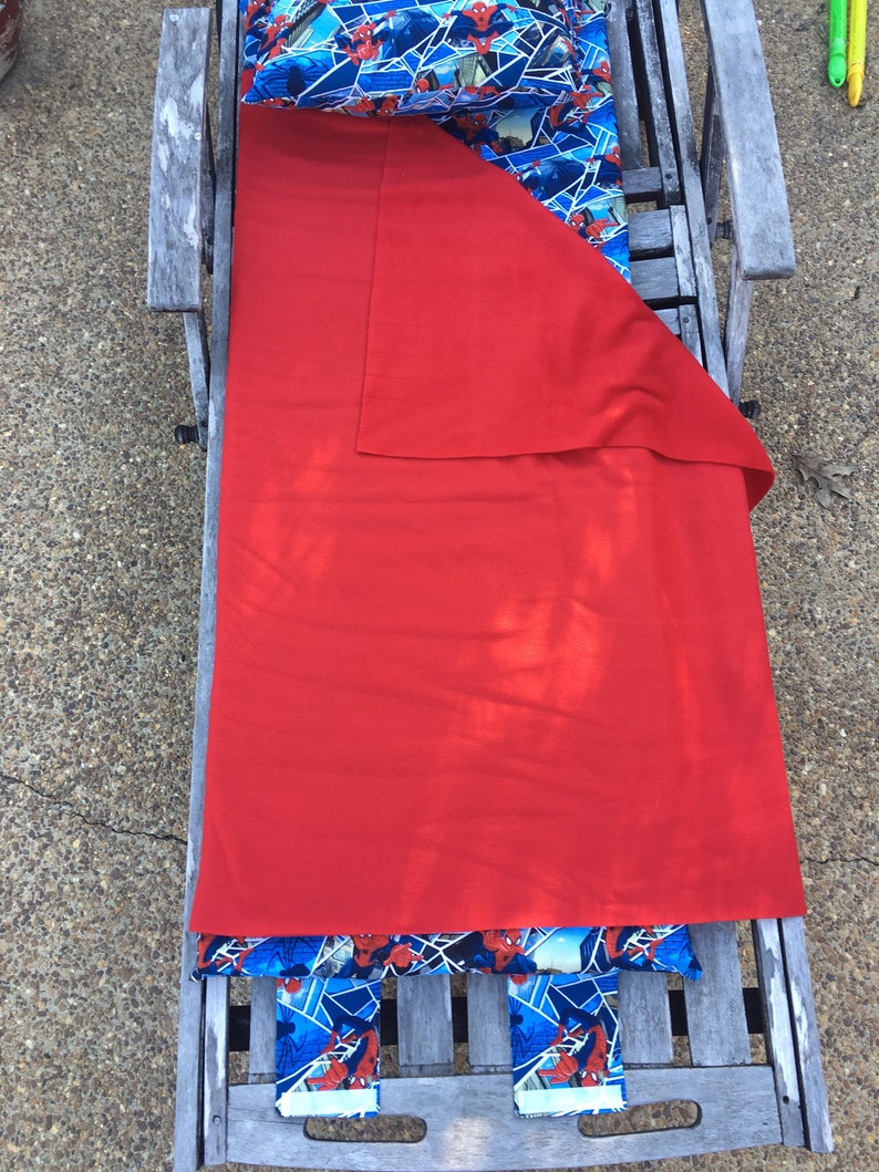 May include: A red sleeping bag with a Spiderman themed pillow and blanket. The sleeping bag is folded and placed on a wooden bench.