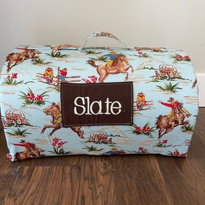 May include: A blue and brown fabric sleeping mat with a cowboy theme. The mat has a handle and is rolled up. The mat has a brown patch with the name "Slate" embroidered on it.