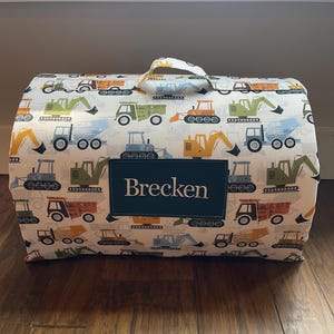 May include: A white fabric storage bin with a handle, decorated with colorful illustrations of construction vehicles. The name "Brecken" is displayed on a dark green rectangular patch on the front of the bin.
