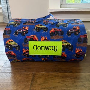 May include: A rolled fabric sleeping mat featuring a blue background with a repeating pattern of monster trucks and orange flames. A rectangular lime green patch is stitched to the center, displaying the name Conway in black cursive text.