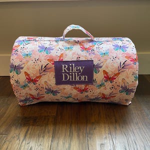 May include: A rolled-up, pink butterfly-themed blanket with a handle. The blanket features a repeating pattern of butterflies and flowers in shades of pink, purple, and blue. A purple patch with the name "Riley Dillon" is sewn on the front.