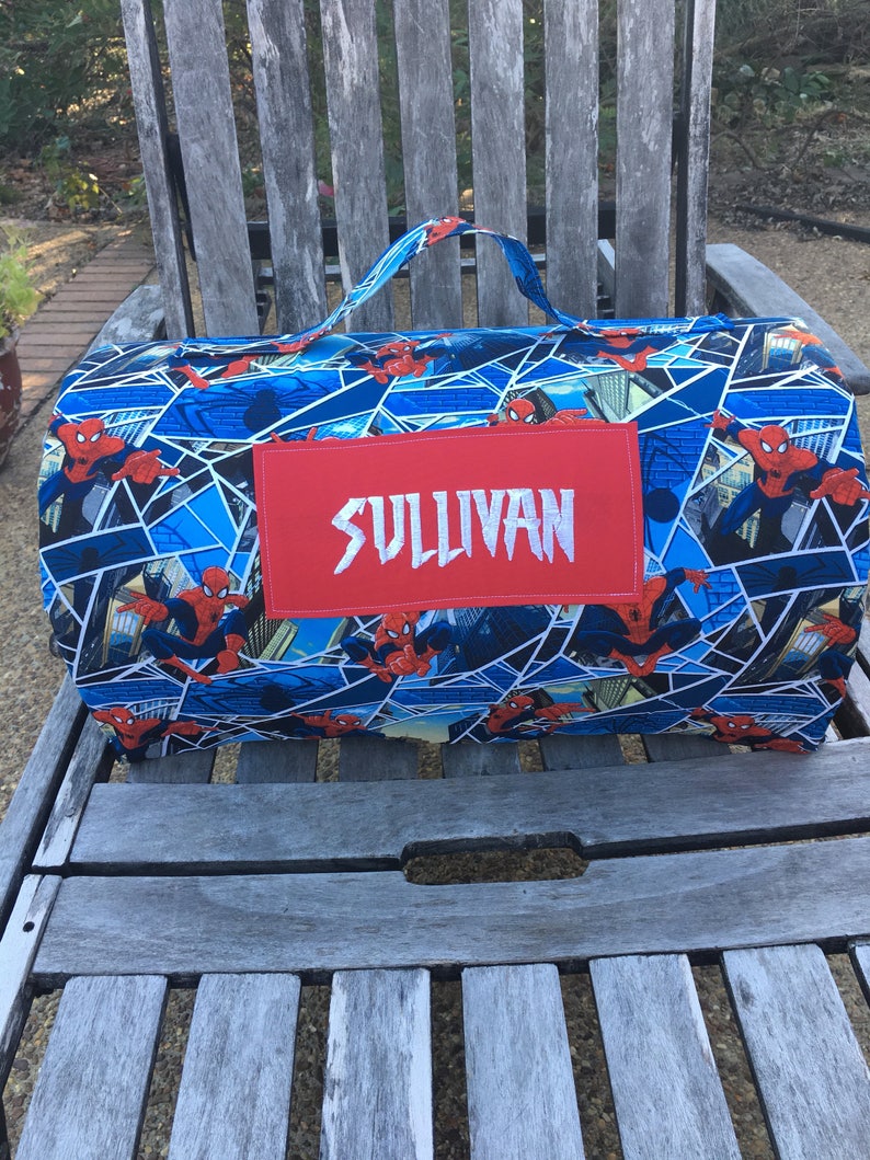 May include: A blue and red sleeping bag with a Spiderman pattern. The sleeping bag has a red name tag with the name "SULLIVAN" embroidered in white.