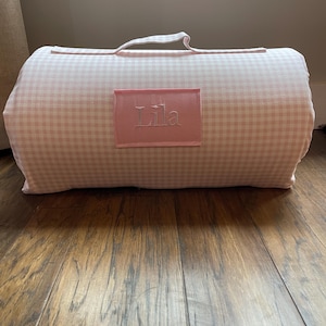 May include: A rolled-up pink and white gingham blanket with a handle. The blanket has a rectangular patch embroidered with the name "Lila" in pink thread. The blanket is resting on a wooden floor.