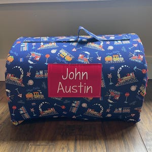 May include: A blue, rectangular, padded item with a handle, featuring a repeating train pattern. A red rectangular patch displays the name "John Austin" in white. The train pattern includes steam trains, stop signs, and the words "Choo Choo".