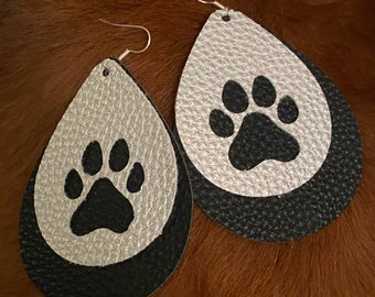 Puede incluir: Un par de pendientes colgantes con un diseño de lágrima de cuero plateado y negro. Cada pendiente presenta una huella de pata negra sobre un fondo plateado.