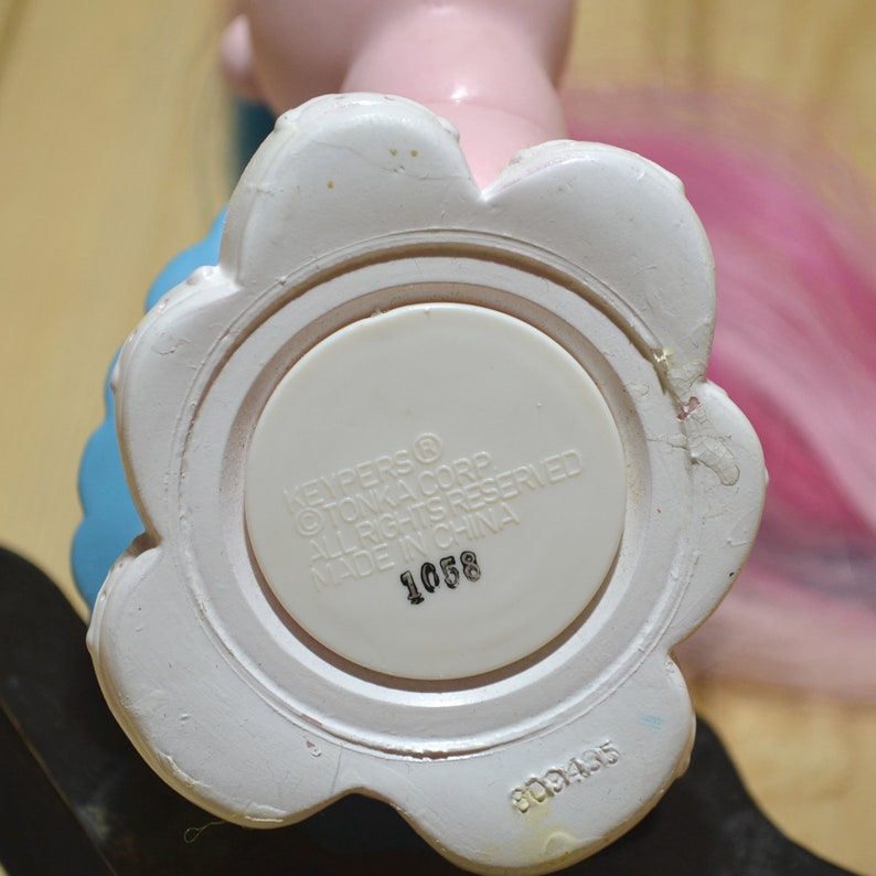 May include: Close-up of the bottom of a pink and blue toy figure. The base is white and flower-shaped, with a circular center. Text on the base reads "KEYPERS &copy; TONKA CORP ALL RIGHTS RESERVED MADE IN CHINA 1058".