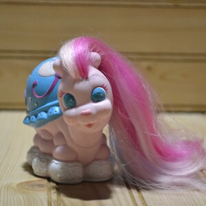 May include: A vintage toy snail with a light pink body, blue shell, and long pink and white hair. The snail has glittery blue eyes and a smiling face. The shell is decorated with stars and a crescent moon.