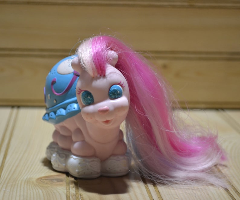 May include: A vintage toy snail with a light pink body, blue shell, and long pink and white hair. The snail has glittery blue eyes and a smiling face. The shell is decorated with stars and a crescent moon.