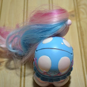 May include: A vintage toy doll with blue and pink hair. The doll's body is light blue with white polka dots and pink accents. The doll has a round shape and a whimsical design, suggesting a collectible or nostalgic item.