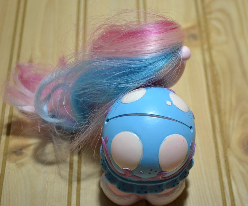 May include: A vintage toy doll with blue and pink hair. The doll's body is light blue with white polka dots and pink accents. The doll has a round shape and a whimsical design, suggesting a collectible or nostalgic item.