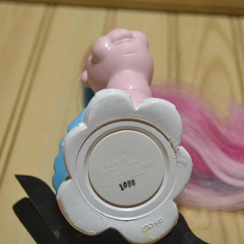 May include: A vintage toy figure with a pink head, blue dress, and pink and purple hair. The white base has a flower-like shape and includes text. The figure is made of plastic.