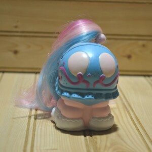 May include: A vintage toy pony with a blue head and pink and blue hair. The pony has large white eyes and pink accents. The body is pink and white. The toy is on a wooden surface.