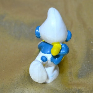 May include: A small, plastic figurine of a Smurf character, viewed from the back. The figure is primarily white with blue accents on the arms, legs, and head. Yellow gloves and blue ear-like details are also visible. The figurine is posed in a crouching position.