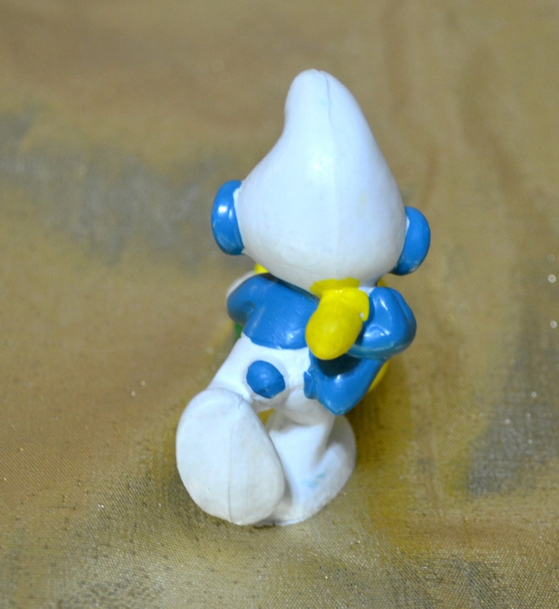 May include: A small, plastic figurine of a Smurf character, viewed from the back. The figure is primarily white with blue accents on the arms, legs, and head. Yellow gloves and blue ear-like details are also visible. The figurine is posed in a crouching position.