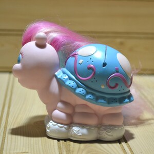 May include: A vintage, light pink toy turtle with a blue shell decorated with white circles and purple swirls. The turtle has a pink and purple mane, blue eyes, and white lace-trimmed feet. A nostalgic collectible toy.