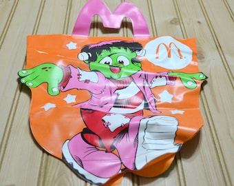 May include: Orange Halloween-themed McDonald's Happy Meal bag featuring a cartoon Frankenstein's monster. The bag has a pink handle and the McDonald's logo. The monster is green with pink clothing and white stars.