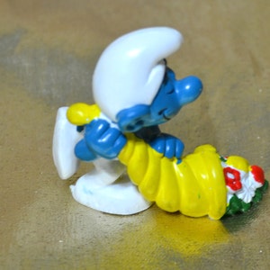 May include: A figurine of a blue Smurf wearing a white hat and yellow outfit, holding a yellow cornucopia filled with red, white, and green flowers. The Smurf is leaning against a white object.