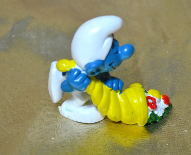 May include: A figurine of a blue Smurf wearing a white hat and yellow outfit, holding a yellow cornucopia filled with red, white, and green flowers. The Smurf is leaning against a white object.