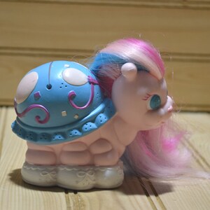 May include: A vintage toy featuring a light pink body and a blue shell adorned with white and purple accents. The toy has a pink, blue, and white mane and tail. The base is white with a decorative design.