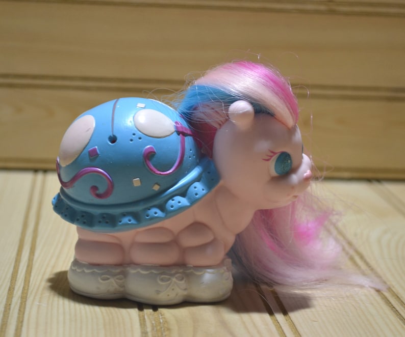 May include: A vintage toy featuring a light pink body and a blue shell adorned with white and purple accents. The toy has a pink, blue, and white mane and tail. The base is white with a decorative design.